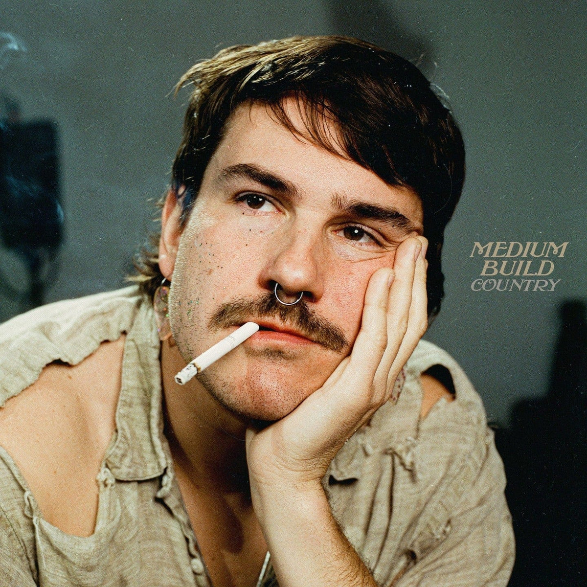 Man with a moustache and cigarette resting his cheek on his hand against a grey-green backdrop