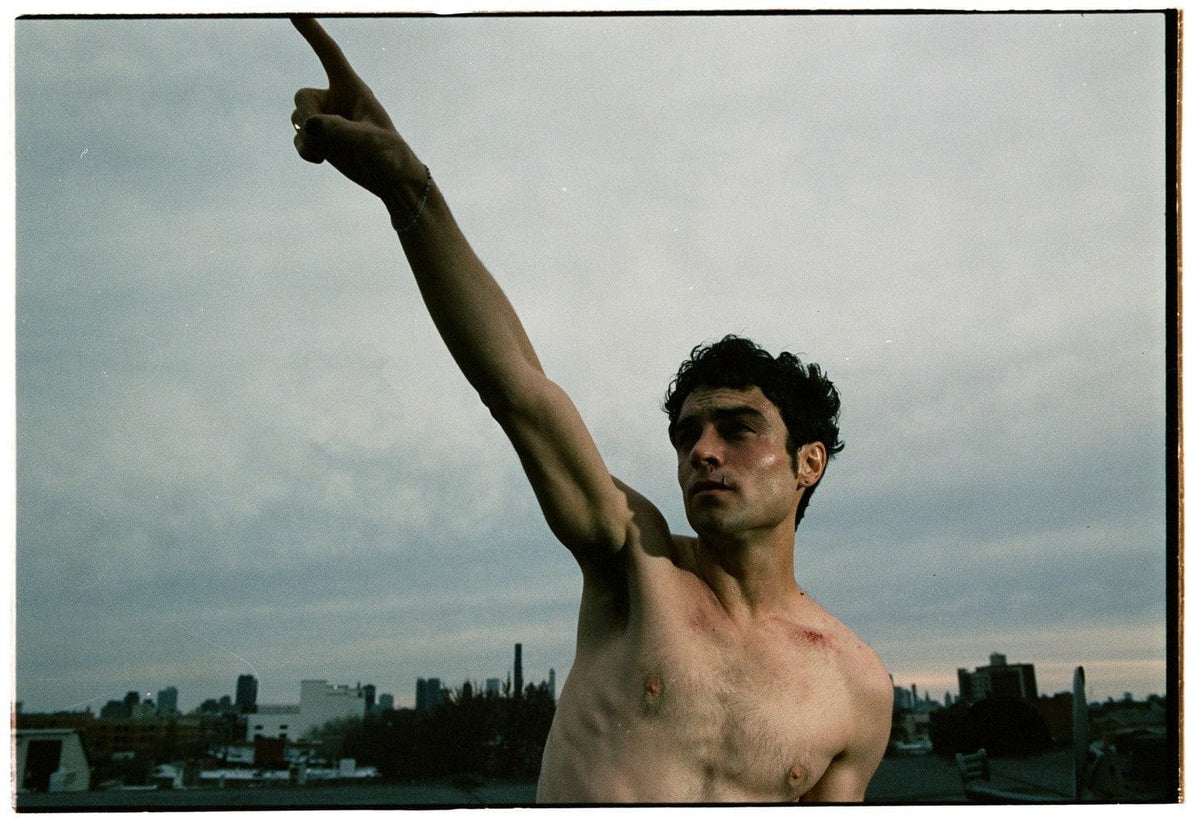 Shirtless man pointing skyward on a rooftop under grey cloud, city skyline behind