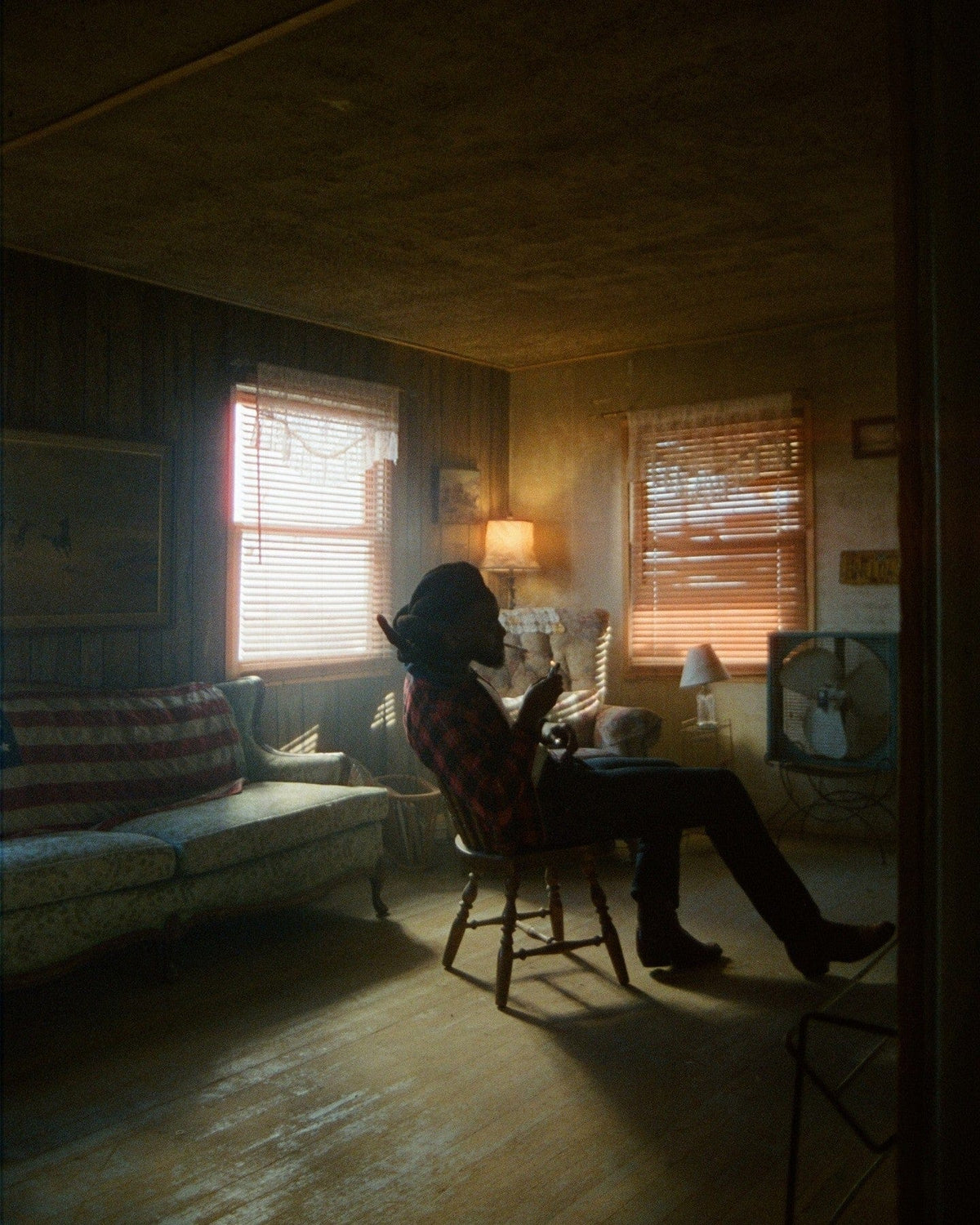 Figure in a wide-brimmed hat sitting in a dim room, light through orange window blinds