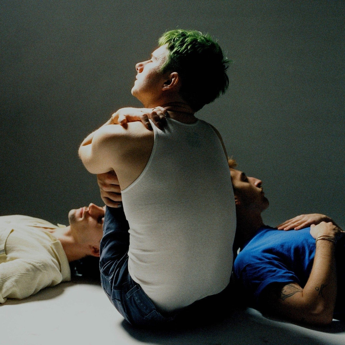 Man with green hair in a white vest seated between two people lying on a studio floor