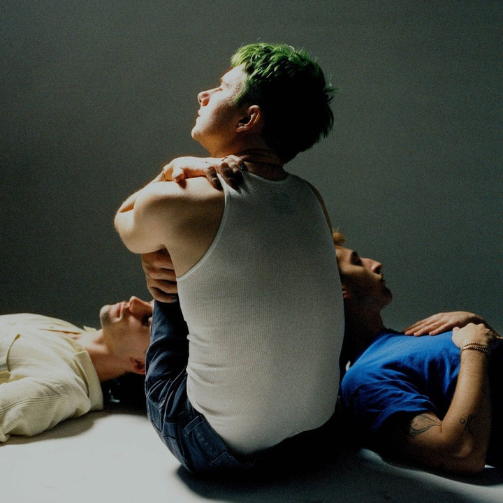 Man with green hair in a white vest seated between two people lying on a studio floor