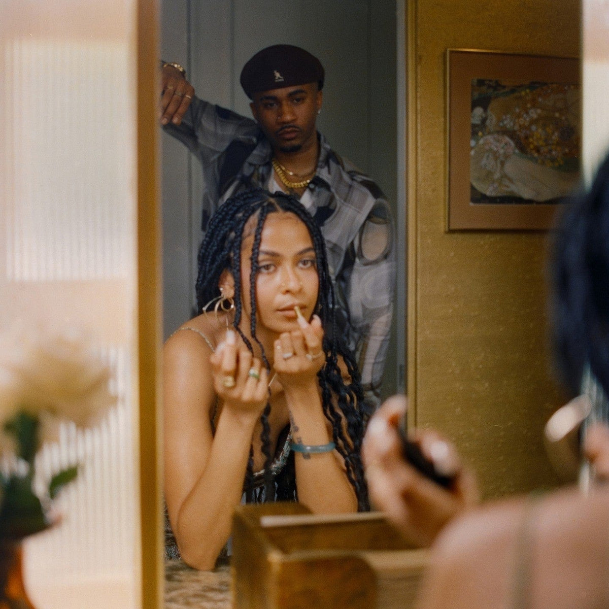 Woman applying lipstick in a mirror, a man in a beret standing behind her