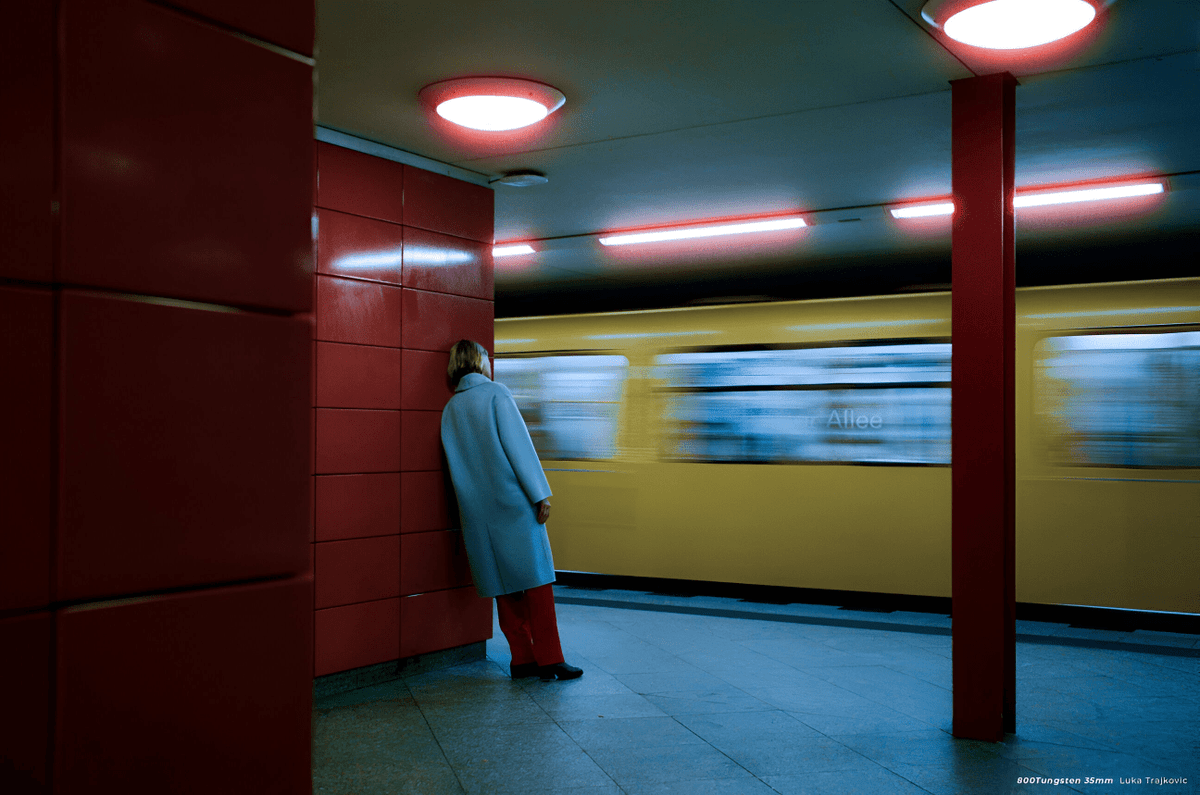 Person in a long pale coat leaning on a red-tiled wall as a yellow train passes in an underground station