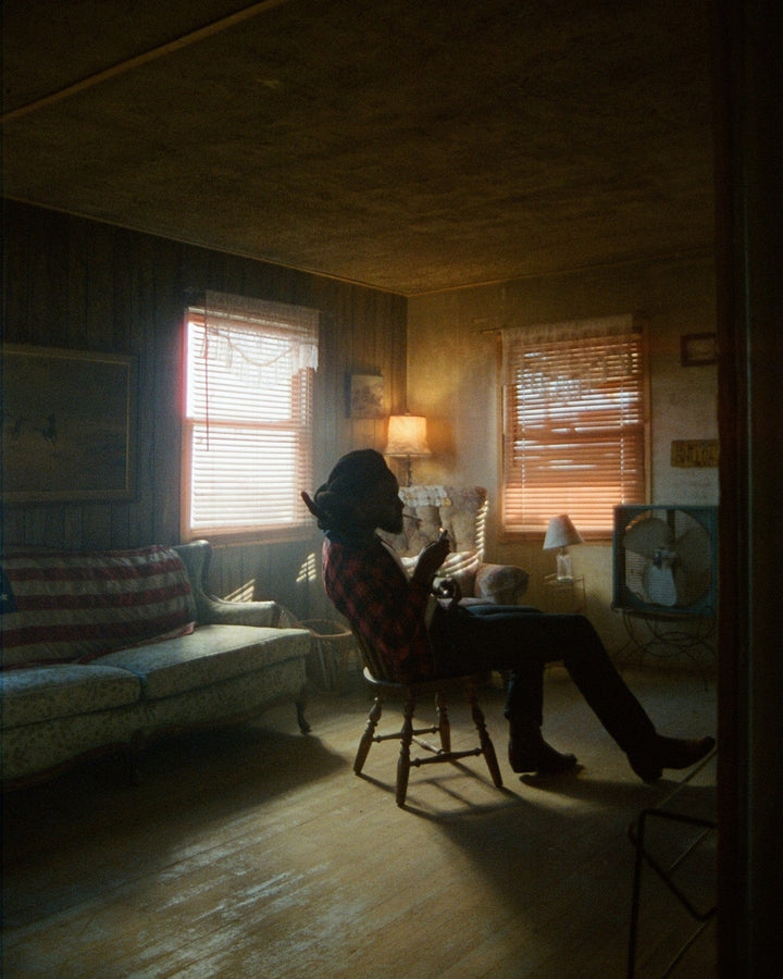 Figure in a wide-brimmed hat sitting in a dim room, light through orange window blinds