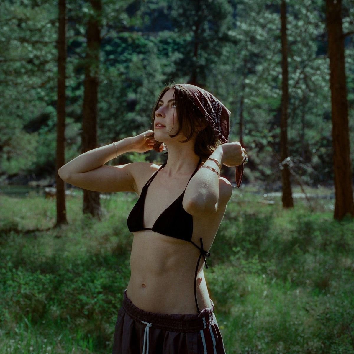 Woman in a headscarf tying back her hair in a sunlit forest clearing