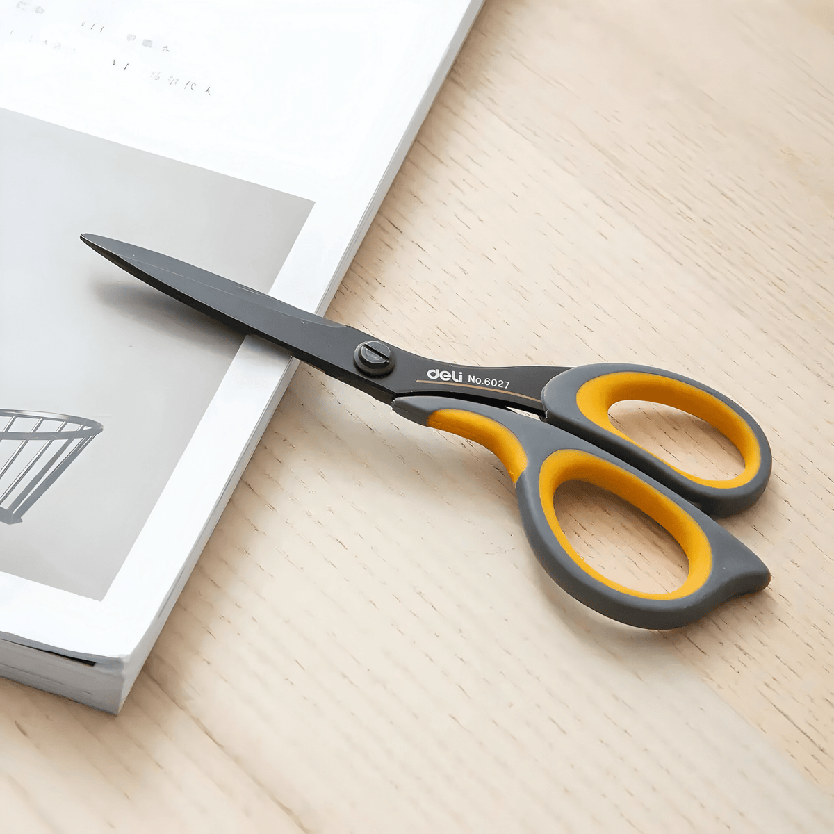 Deli E6027 scissors lying on a wooden desk beside an open magazine