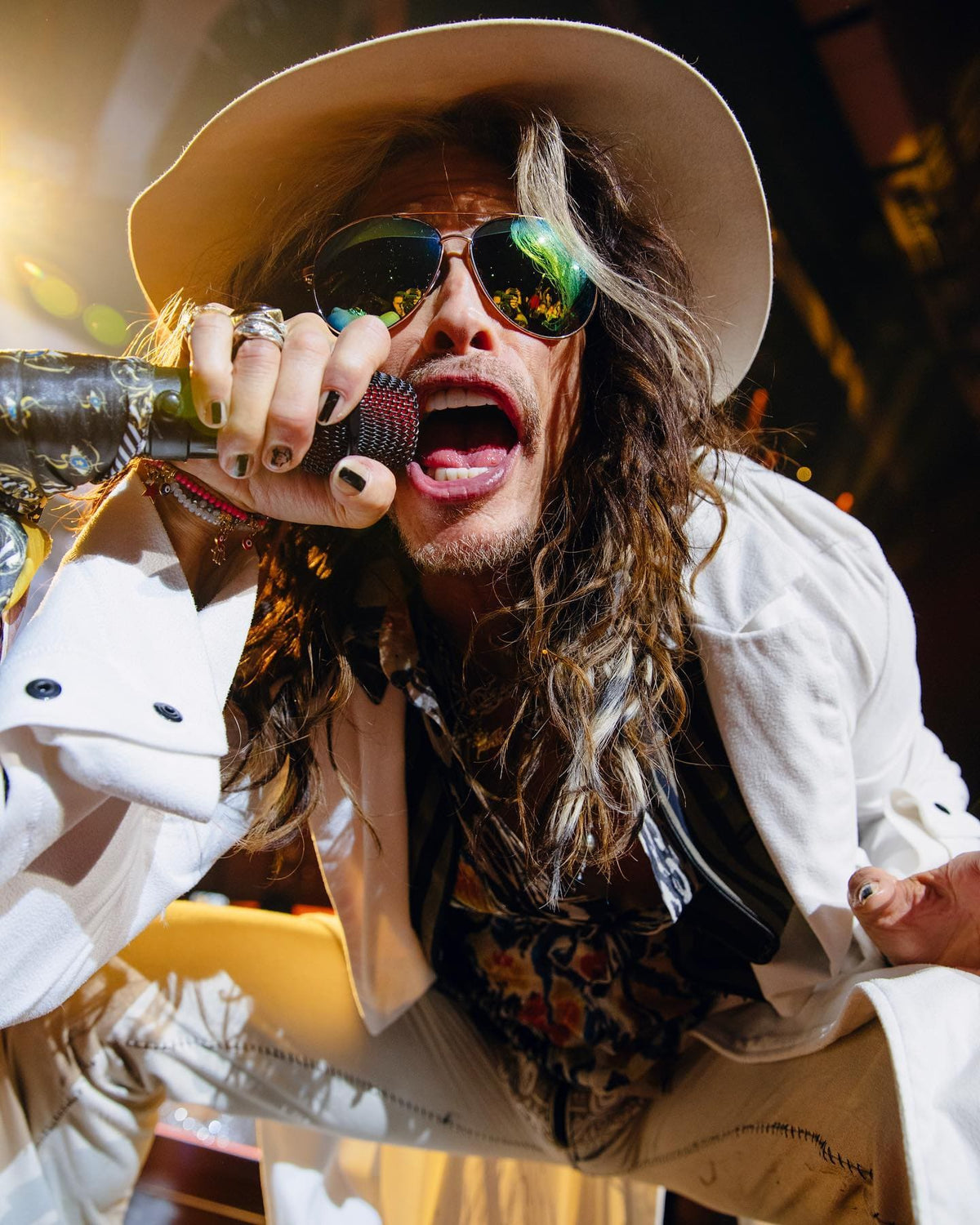 Singer in a white hat and sunglasses performing close to a handheld mic under warm stage lights