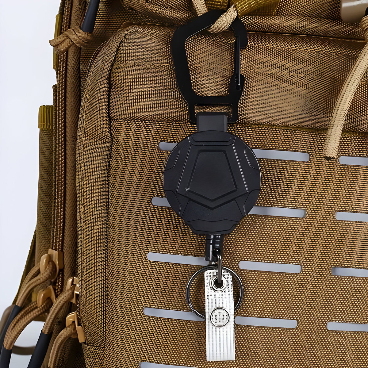 Retractable carabiner reel clipped to a tan backpack's webbing, with key ring and clip strap hanging below