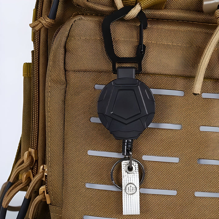 Retractable carabiner reel clipped to a tan backpack's webbing, with key ring and clip strap hanging below